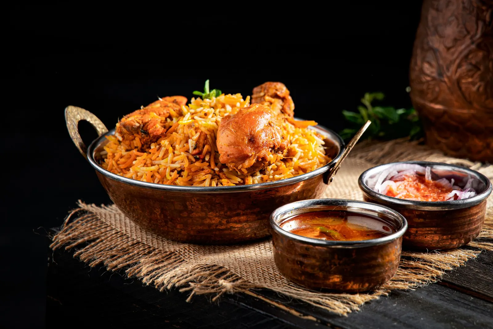 Chicken biryani in a copper kadhai with sides on a dark table