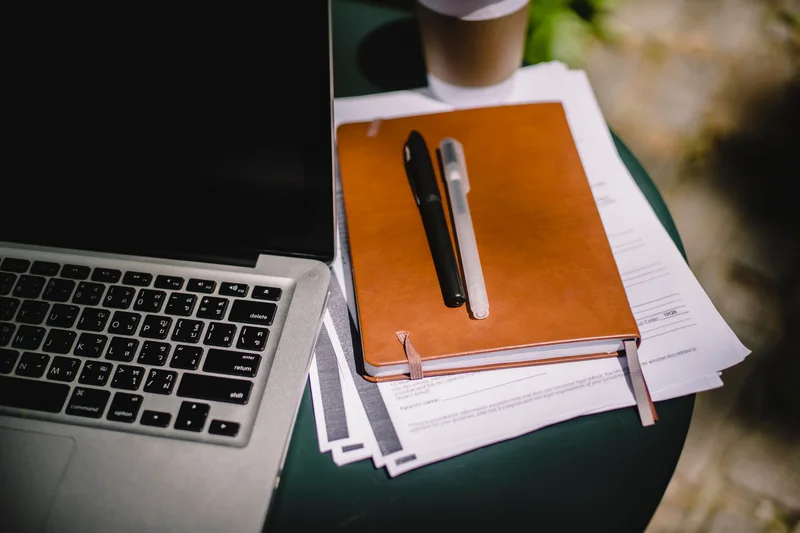 A leather notebook and papers beside a laptop